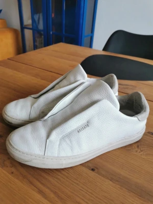 Vita Axel Arigato sneakers i läder - Snygga vita sneakers från Axel Arigato i mjukt läder med clean design och diskret logga på sidan. Modellen har låg profil och bred vit sula, samt en unik ovandel utan synliga snören för en minimalistisk vibe.
