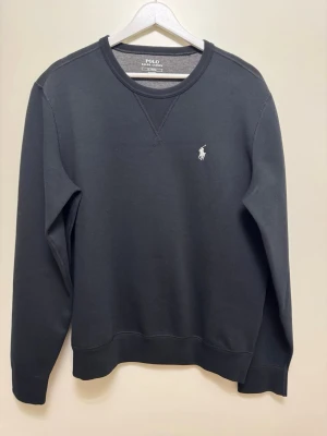 Svart sweatshirt från Polo Ralph Lauren - Som ny! Klassisk svart sweatshirt från Polo Ralph Lauren med rund halsringning och vit broderad logga på bröstet. Tröjan har långa ärmar och ribbade muddar vid ärmslut och nederkant. Perfekt för en stilren och avslappnad look.