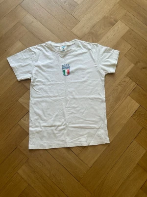 Italia t-shirt  - Säljer en vit t-shirt med Italiens landslagssköld och texten 'ITALIA' tryckt både fram och bak. T-shirten har korta ärmar och är gjord i mjuk bomull. Perfekt för sommaren om du ska till Italien tex annars bara en snygg t-shirt. Köpt i Italien. 