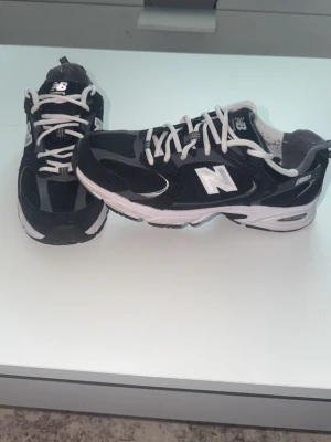 Svarta New Balance 530 - Klassiska svarta New Balance 530 sneakers med vita detaljer och grå inslag. Skorna har en sportig siluett, mesh- och mockadetaljer samt en vit sula med grå partier. Perfekta för en streetwear-look och riktigt bekväma att gå i.