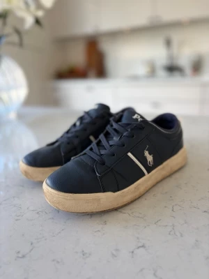 Marinblå Polo Ralph Lauren sneakers - Snygga marinblå sneakers från Polo Ralph Lauren med klassisk vit logga på sidan och vita detaljer. Skorna har lågt skaft, rund tå och ovandel i läderimitation. Sparsamt använda. 