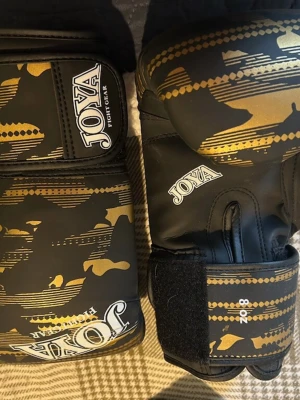 Svarta boxningshandskar från Joya barn  - Säljer ett par svarta boxningshandskar från Joya med guldfärgade kamouflagemönster och vita logotyper. Handskarna har kardborreband för enkel justering och sitter stabilt på handen. Perfekta för träning inom kampsport eller boxning. 8 Oz 