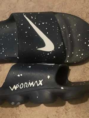 Nike Vapormax svarta sandaler - Svarta Nike Vapormax sandaler med vitt stänkmönster och stor vit Swoosh-logga på ovandelen. Tillverkade i syntetmaterial med en chunky sula för extra komfort. Perfekta för chill dagar och streetwear-stil.