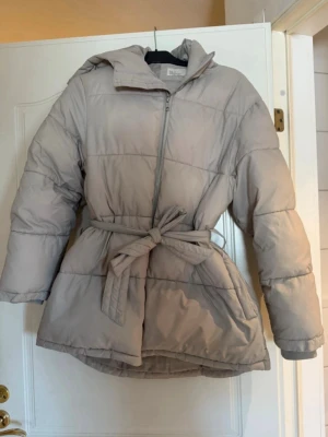 Beige pufferjacka med bälte och huva - Snygg beige pufferjacka från Nelly med avtagbart bälte i midjan och huva. Jackan har dragkedja framtill, två fickor och quiltad design. Perfekt för kalla dagar när du vill hålla dig varm och samtidigt se trendig ut. Stor i storleken. Innerfickan har blivit missfärgad av telefon men inget som syns när man har på sig den 