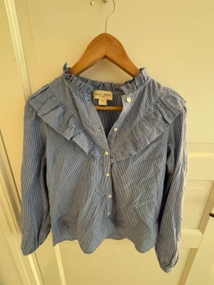 Blårandig blus med volang från Holly & White - Snygg blårandig blus från Holly & White med vita knappar och volangdetalj längs kragen och axlarna. Blusen har lång ärm och klassisk krage.