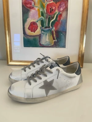 Golden Goose sneakers - Snygga vita sneakers från Golden Goose med grå stjärna på sidan, gråa skosnören och mörkgrå häl. Klassisk låg modell med slitna detaljer och svart sula undertill. Perfekta för en trendig och avslappnad look.