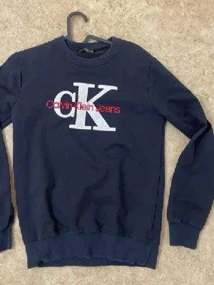 Mörkblå sweatshirt från Calvin Klein - Mörkblå sweatshirt med stor vit CK-logga och röd Calvin Klein Jeans-text på bröstet. Klassisk rund halsringning och ribbade muddar vid ärmslut och nederkant. Perfekt för en avslappnad och stilren look.
