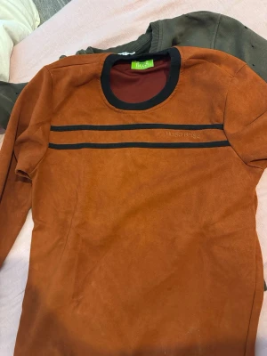 Rostbrun tröja från Hugo Boss - Snygg rostbrun långärmad tröja från Hugo Boss med svarta detaljer och två svarta ränder över bröstet. Rund halsringning med svart kant och diskret broderad logga. Perfekt för en stilren och avslappnad look.