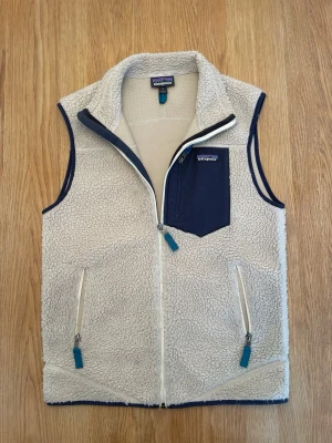 Patagonia väst - Beige fleeceväst från Patagonia med marinblå detaljer och dragkedjor. Hög krage, två fickor framtill och en bröstficka med dragkedja. Relativt ny väst med inga defekter. Bra skick och strl S.