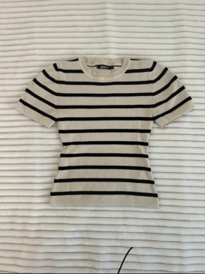 Randig beige topp från Gina Tricot - Snygg beige topp från Gina Tricot med svarta horisontella ränder. Modellen är figurnära med korta ärmar och rund halsringning. Materialet är ribbat, vilket ger en skön känsla och en stilren look.