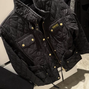Svart quiltad jacka från Barbour - Svart quiltad jacka från Barbour med guldiga knappar, dragkedja och flera fickor. Klassisk design med markerad midja och hög krage. Perfekt för dig som gillar stilren och tidlös look. Barbour jackan är en märkes jacka värd 4000kr men Säljer för 2000 men ni kan komma med rimliga förslag.