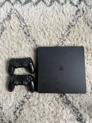 PlayStation 4 Slim med två kontroller - Säljer en svart PlayStation 4 Slim från Sony tillsammans med två matchande DualShock 4-kontroller. Konsolen har en stilren, matt finish och klassisk fyrkantig form. Perfekt för gamingkvällar och multiplayer med vänner.