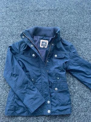 Mörkblå vindjacka från Staccato och 152cm - Snygg mörkblå vindjacka från Staccato med flera fickor och dragkedja framtill. Jackan har hög krage, tryckknappar och detaljer i metall. Materialet är lätt och vindtätt, perfekt för blåsiga dagar.