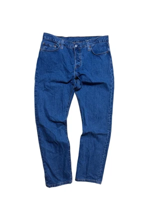 Blå Levi's 501 raka jeans - Klassiska blå Levi's 501 jeans med rak passform och fem fickor. Jeansen har knappgylf och tydliga sömmar, tillverkade i 100% bomull. Bakfickorna har den ikoniska V-formade sömmen och röd Levi's-lapp bak i midjan.