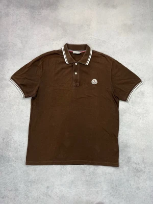 Brun pikétröja från Moncler - Snygg brun pikétröja från Moncler med klassisk krage och vita ränder på krage och ärmslut. Tröjan har knappslå framtill och Moncler-logga broderad på bröstet. Perfekt för en clean och stilren look.
