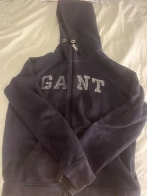 Mörkblå hoodie från GANT - Snygg mörkblå hoodie från GANT med dragkedja och stor huva. Framtill finns ett stort GANT-tryck i vitt. Hoodien har fickor på sidorna och ribbade muddar vid ärmslut och nederkant. Perfekt för en avslappnad och cool stil.