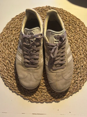 Adidas Gazelle grå sneakers - Säljer ett par klassiska Adidas Gazelle sneakers i grå mocka med vita detaljer och de ikoniska tre ränderna på sidan. Skorna har lågt skaft, grå snörning och en beige sula. Perfekta för dig som gillar retrostil och streetwear.