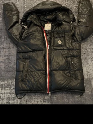 Svart dunjacka från Moncler - Snygg svart dunjacka från Moncler med glansig finish, huva och dragkedja med röd, vit och blå detalj. Jackan har två snedställda fickor med dragkedja och en bröstficka med lock. Moncler-logga på bröstet. Perfekt för kalla vinterdagar.storlek xl passar s/m