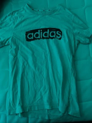 Vit t-shirt med Adidas-logga - Säljer en vit t-shirt från Adidas med stor svart logga tryckt på bröstet. Klassisk rund hals och korta ärmar. Tillverkad i mjukt bomullsmaterial som är skönt mot huden. Perfekt för en sportig och avslappnad stil.