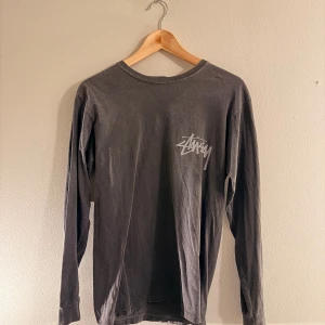 Grå långärmad Stüssy t-shirt - Säljer en grå långärmad t-shirt från Stüssy med deras ikoniska logga tryckt i vitt på bröstet. Tröjan har en klassisk rund halsringning och är gjord i mjuk bomull som känns skön mot huden. Perfekt för en avslappnad streetwear-look.