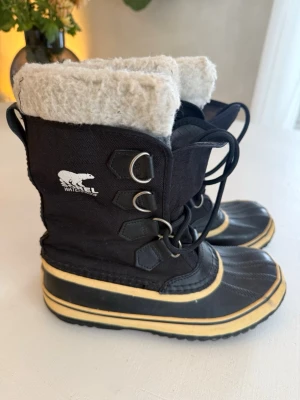 Svarta vinterstövlar från Sorel - Säljer ett par svarta vinterstövlar från Sorel med vattenavvisande material, snörning och varm, beige fuskpälsfodring. Stövlarna har robust gummisula och detaljer i gult och svart. Perfekta för kalla dagar och snöiga äventyr.