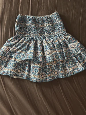 Blå mönstrad kortkjol Meet x Tiger - Supersöt kortkjol från Meet me there! blått med paisley-inspirerat mönster. Kjolen har bred smock i midjan för skön passform och är gjord i ett lätt bomullstyg. Perfekt för dig som gillar färg och mönster! Stretchig passar xxs-s (inbyggda shorts) 