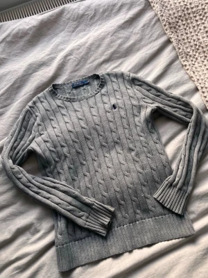 Grå kabelstickad tröja Polo Ralph Lauren - Snygg grå kabelstickad tröja från Polo Ralph Lauren med rund halsringning och diskret broderad logga på bröstet. Tröjan har långa ärmar och ribbade muddar vid ärmslut och nederkant. Perfekt för lager-på-lager och klassisk stil.