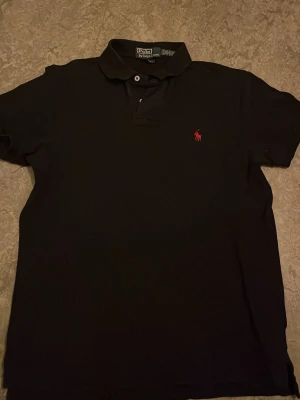 Svart pikétröja från Polo Ralph Lauren - Klassisk svart pikétröja från Polo Ralph Lauren med röd broderad logga på bröstet. Slim fit-modell med korta ärmar och knappar vid kragen. Tillverkad i mjuk bomull som känns skön mot huden.