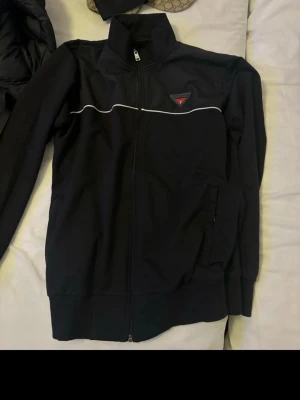 Svart zip hoodie från Guess - Snygg svart hoodie från Guess med dragkedja framtill och vit piping över bröstet. Guess-logga i rött och vitt på vänster sida. Mjuk bomullsblandning och två fickor framtill. Perfekt för en casual och trendig look.