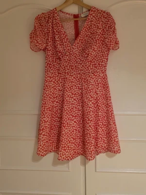 Röd blommig kortklänning med v-ringning - Söt röd kortklänning med vitt blommönster och v-ringad hals. Klänningen har korta ärmar och en lätt utställd kjol.