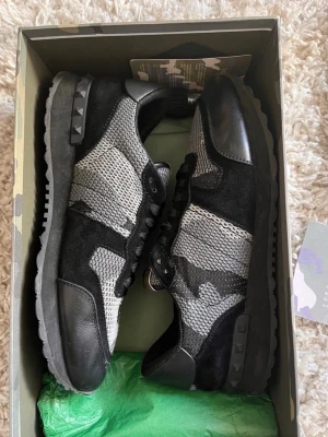 Valentino svart/grå sneakers - Snygga sneakers från Valentino i svart och grått med camomönster. Skorna har en mix av mocka, mesh och läderdetaljer samt nitar på sulan. Perfekta för dig som vill ha en unik och trendig look. Levereras med originalkartong och tillbehör. Skorna är använda sparsamt och i mycket bra skick. Om du har någon fråga är det bara att höra av sig mvh Levi!