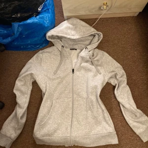 Ljusgrå kofta - Säljer en ljusgrå hoodie med dragkedja framtill och stor huva. Tröjan har två fickor på sidorna och breda muddar vid ärmslut och nederkant. Perfekt för en avslappnad stil och enkel att matcha med det mesta.