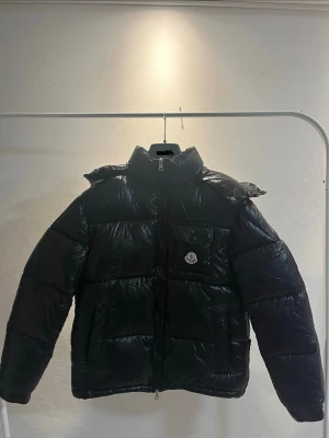 Moncler original jacka – storlek 3  - Snygg svart dunjacka från Moncler med glansig finish, hög krage och huva. Jackan har dragkedja framtill, fickor på sidorna och en bröstficka med Moncler-logga. Perfekt för kalla vinterdagar och riktigt trendig look. jackan är köpt av mig. Alla bilder är tagna av mig och visar detaljer, etiketter och certilogo. Jackan är i gott skick. Man kan är ta av luvan och sätta på. Men passar även storlek 2