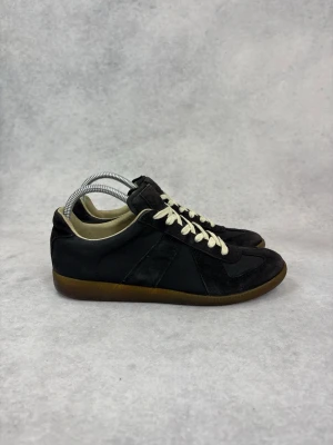 Svarta sneakers med mockadetaljer - Stilrena svarta sneakers med vita snören och bruna gummisulor. Ovandelen är i en mix av mocka och textil, vilket ger en schysst kontrast. Låg profil och klassisk siluett som funkar till det mesta. Perfekta för dig som gillar en clean och tidlös look.