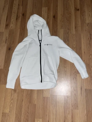 Vit hoodie från Sail racing - Snygg vit hoodie från sail Racing med svart dragkedja och två stora fickor framtill. Hoodien har en klassisk huva och stilren logga på bröstet. Perfekt för dig som gillar enkel och sportig stil. Materialet känns mjukt och bekvämt mot huden.