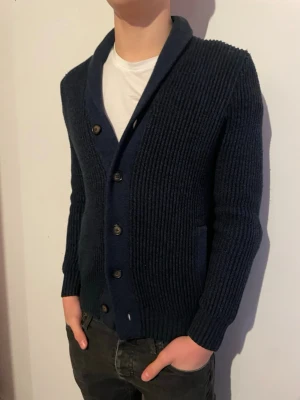 Mörkblå stickad cardigan från marks & Spencer  - Snygg mörkblå stickad kofta med sjalkrage och knäppning framtill. Koftan har ribbad struktur, två fickor och en klassisk, avslappnad passform. Perfekt att slänga över en t-shirt för en chill look.