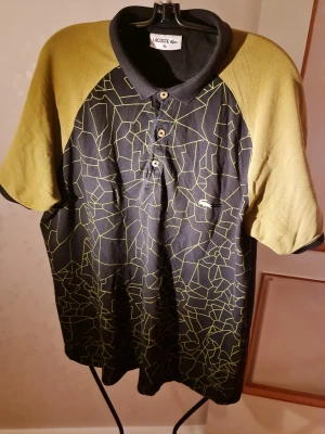 Svart och gul mönstrad piké Lacoste - Cool pikétröja från Lacoste i svart med gula ärmar och grafiskt mönster framtill. Klassisk krage, tre knappar och diskret Lacoste-logga på bröstet. Perfekt för dig som vill sticka ut med en unik topp.