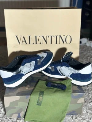 Blå och vita valentino rockrunners  - Snygga sneakers från Valentino i blått, vitt och grått med meshdetaljer och mocka. Skorna har en sportig siluett med vita sulor och mörkblå detaljer. Perfekta för dig som vill ha en exklusiv och trendig look. Helt nya direkt ifrån butik allt og ingår