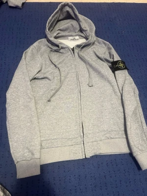 Grå zip hoodie från Stone Island - Snygg gråmelerad hoodie från Stone Island med dragkedja framtill och klassisk logopatch på vänster ärm. Tröjan har huva med dragsko och ribbade muddar vid ärmslut och nederkant. 