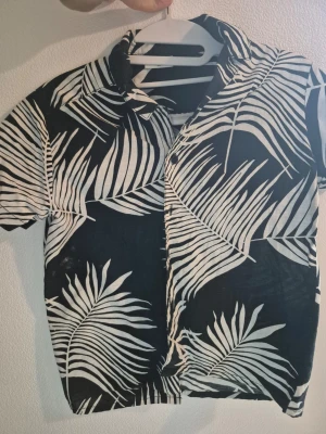 Barn Svart kortärmad skjorta med palmblad stl 134 - Snygg svart skjorta med vita palmbladsmönster. Skjortan har korta ärmar, klassisk krage och knäppning framtill. Perfekt för en avslappnad och trendig stil.