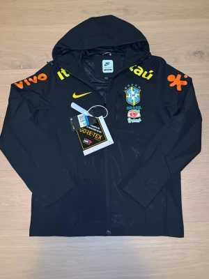 Nike Brazil regnjacka GORE-TEX svart - Svart regnjacka från Nike med Brazil-logga och GORE-TEX-material. Jackan har huva, dragkedja framtill och tryck i gult och orange på bröst och ärmar. Perfekt för dig som gillar sportig stil och vill hålla dig torr i regnet.