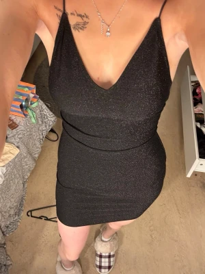 Glittrig svart bodycon klänning - Snygg svart klänning med glittereffekt och smala axelband. Djup v-ringning framtill och öppen rygg med tunna band. Figurnära passform och kort längd, perfekt för en trendig look.