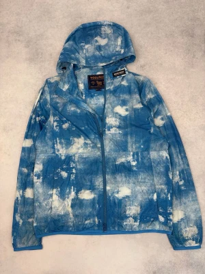 Blå mönstrad vindjacka från Woolrich - Snygg blå vindjacka från Woolrich med vitt molnliknande mönster. Jackan har huva, hel dragkedja och är tillverkad i lätt syntetmaterial. Perfekt för dig som vill sticka ut med en unik look.