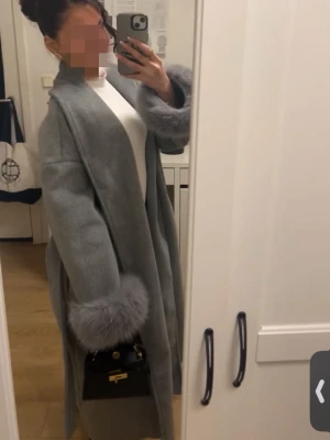 Ljusgrå lång kappa med fuskpäls - Snygg ljusgrå kappa med långa ärmar och breda muddar i fuskpäls. Kappan har en rak och avslappnad passform utan synliga knappar eller dragkedja. Är själv 158cm och den är lång på mig.