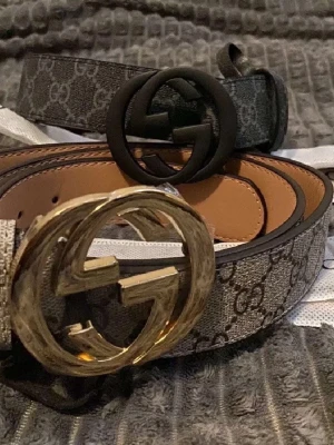 Gucci bälte med GG-spänne - Säljer två Gucci-bälten med klassiskt GG-mönster och stora GG-spännen, ett i guld och ett i svart. Bältena är i beige och grå toner med det ikoniska monogramtrycket. Perfekt accessoar för att lyfta vilken outfit som helst.