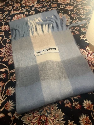 Blå-rutig halsduk från Acne Studios - Mjuk och fluffig halsduk från Acne Studios i en ljusblå och grå-rutig design med inslag av vitt. Halsduken har fransar i ändarna och en tydlig Acne Studios-logga. Perfekt accessoar för att lyfta din outfit med en stilren touch.