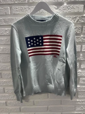Ljusblå stickad tröja från Ralph Lauren - Stickad ljusblå tröja från Ralph Lauren med amerikansk flagga i rött, vitt och blått på bröstet samt RL-broderi. Tröjan har rund halsringning, långa ärmar och ribbade muddar. Perfekt för dig som gillar klassisk och preppy stil.