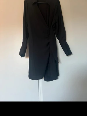 Svart omlottklänning med långa ärmar - Stilren svart omlottklänning med långa ärmar och v-ringad hals. Klänningen har en knytning i midjan som ger en smickrande siluett och en kortare längd. Perfekt för dig som gillar minimalistisk och elegant stil.