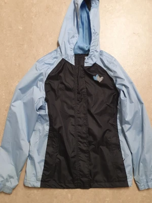 Vindjacka - Snygg vindjacka i ljusblått och svart med huva och dragkedja. Jackan har kardborrestängning över dragkedjan och ett litet hjärtmärke på bröstet. Tillverkad i tunt, vindtätt material som passar perfekt för blåsiga dagar.