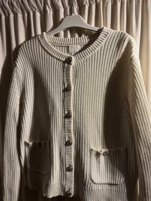 Beige ribbstickad kofta med guldknappar  - Säljer en beige ribbstickad kofta med rund halsringning och dekorativa metallknappar i guld framtill. Perfekt för lager på lager och enkel att matcha till olika outfits. Sparsamt använd